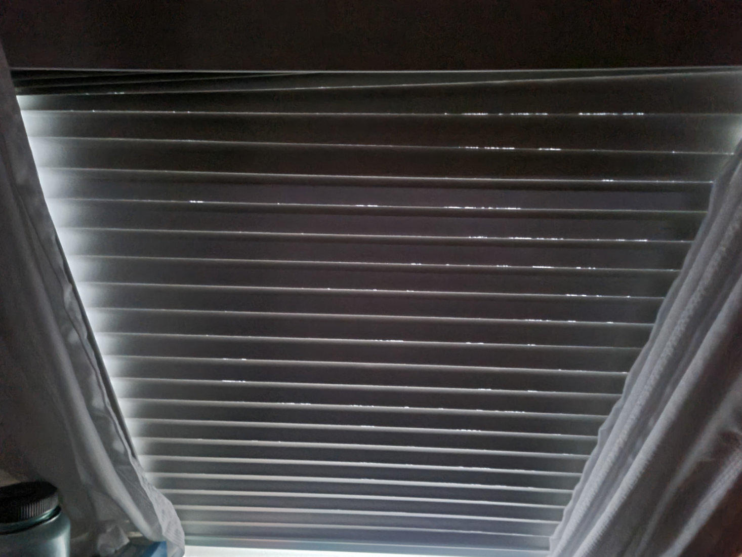 Innenansicht eines geschlossenen Rollladens mit horizontalen Lamellen, links und rechts Vorh&auml;nge, oben schmaler Lichtspalt, unten links zylindrischer Beh&auml;lter