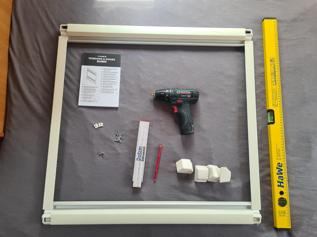 Wei&szlig;er rechteckiger Metallrahmen, Bedienungsanleitung, schwarzer und roter Akkuschrauber, mehrere Schrauben und kleine Metallwinkel, drei wei&szlig;e Kunststoffkappen, roter Stift, gelbe Wasserwaage auf grauem Stoff