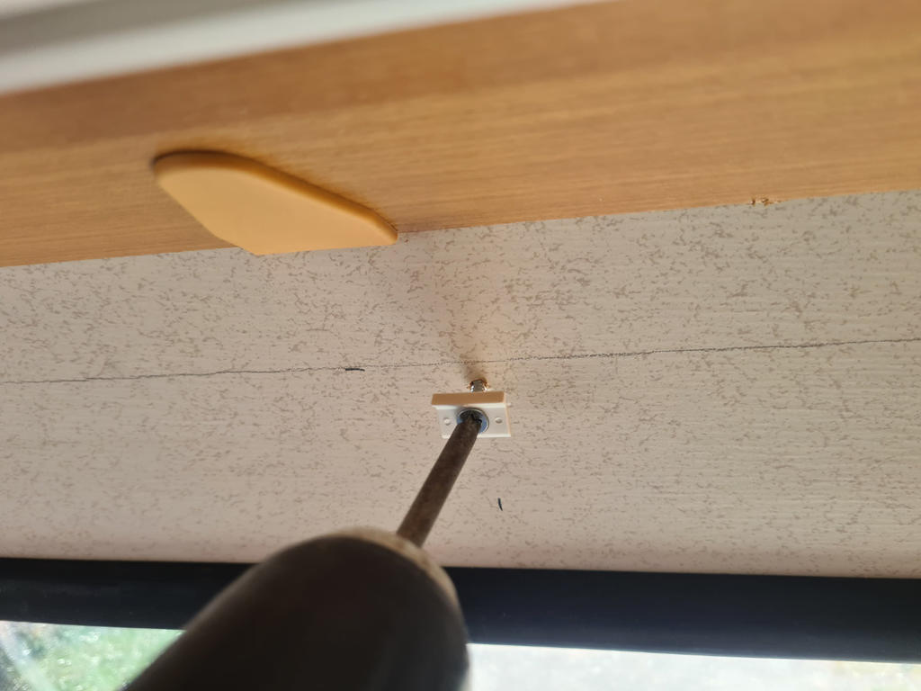 Schwarzer Akkuschrauber mit Bit an einer Schraube in einer kleinen wei&szlig;en Platte an einer hellen Deckenplatte; dar&uuml;ber Holzleiste mit gelbem Keil, unten Fensterrahmen und Au&szlig;envegetation