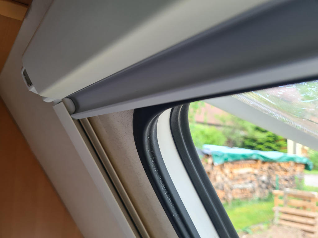 Teilweise ge&ouml;ffnete Fensterklappe mit sichtbarem Rollo und Fensterrahmen; au&szlig;en gestapelte Holzscheite unter gr&uuml;ner Plane, Gras und Str&auml;ucher