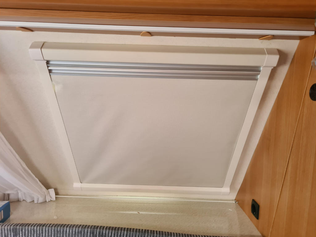 Wei&szlig;es heruntergezogenes Rollo in rechteckigem Holzfensterrahmen, leichter Vorhang links, Fensterbank mit kleinen Gegenst&auml;nden, Steckdose rechts