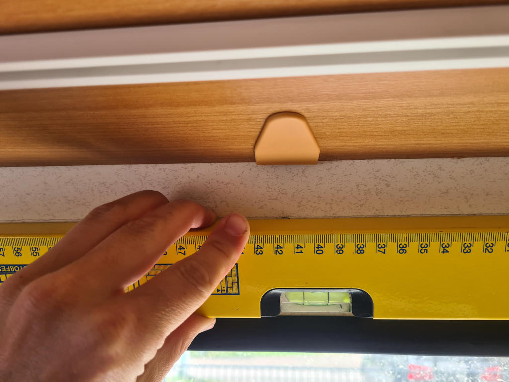 Hand h&auml;lt eine gelbe Wasserwaage an einem hellen Holzrahmen; oberhalb ein kleiner beiger Kunststoffclip, unten Teil eines Fensters mit unscharfer Au&szlig;enansicht