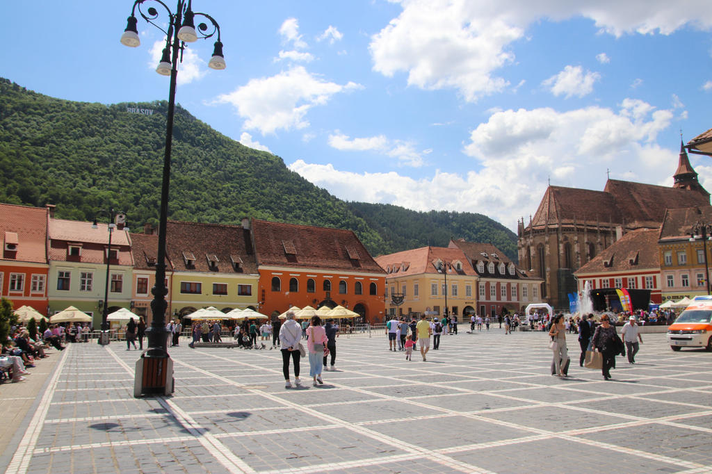 Gro&szlig;er gepflasterter Marktplatz mit Menschen, bunten H&auml;usern, einer Stra&szlig;enlaterne, Kirche, Bergen im Hintergrund und blauem Himmel