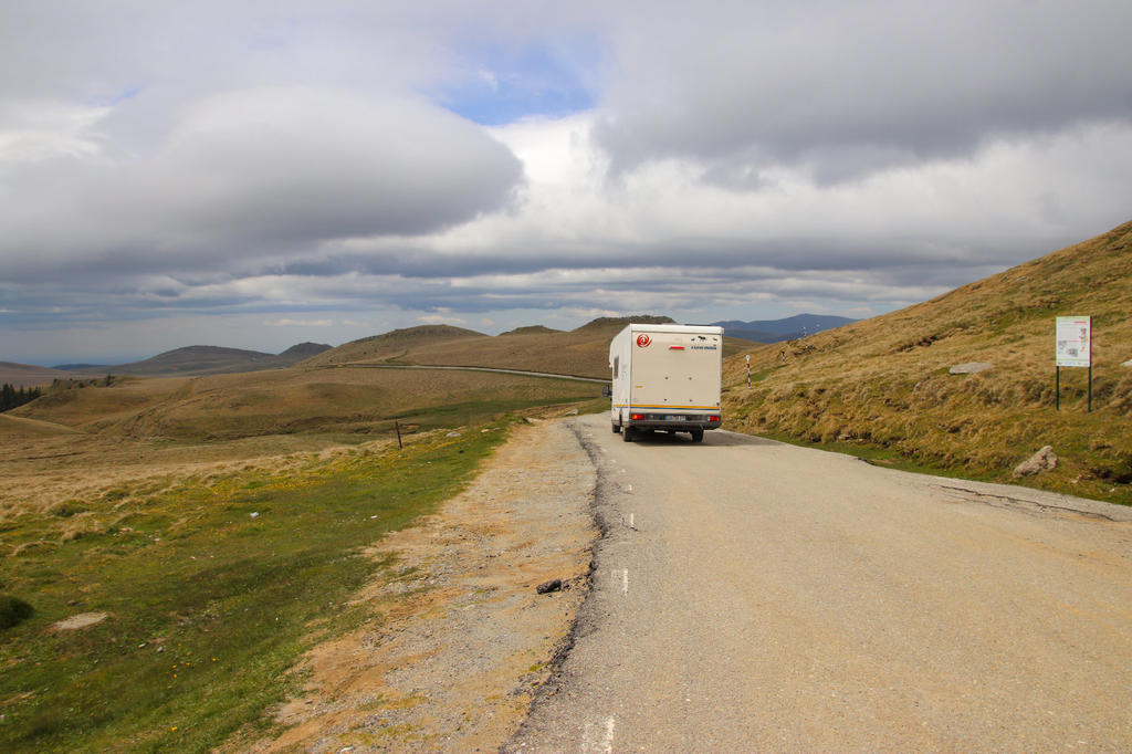 R&uuml;ckansicht eines Wohnmobils auf einer Landstra&szlig;e in h&uuml;geliger Graslandschaft unter bew&ouml;lktem Himmel