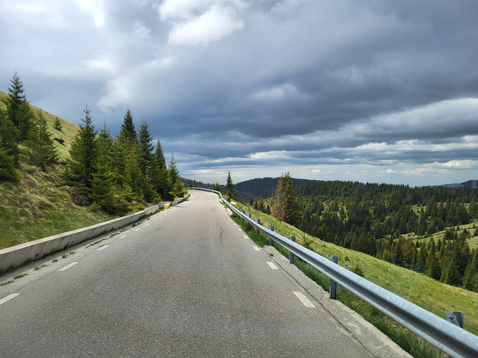 Asphaltierte Stra&szlig;e in Linkskurve, Leitplanke rechts, Nadelb&auml;ume links, grasbewachsene H&auml;nge und dunkle Wolken am Himmel
