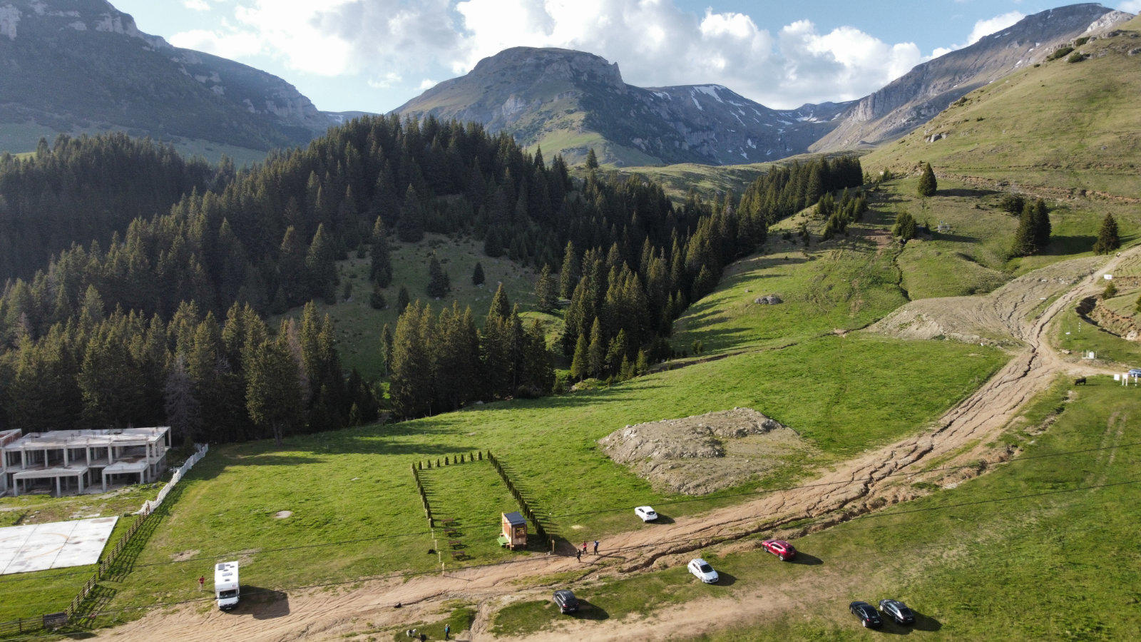 Bergtal mit gr&uuml;nen Wiesen und Nadelwald, Gipfel im Hintergrund, wolkiger Himmel, unbefestigte Fahrspuren mit mehreren Fahrzeugen, ein wei&szlig;es Geb&auml;ude links, Erdhaufen und kleine Z&auml;une auf der Wiese