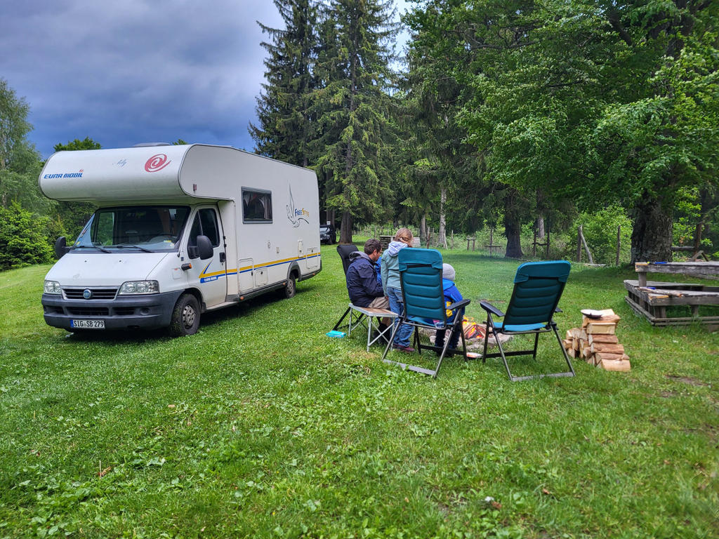 Wei&szlig;es Wohnmobil auf einer Wiese; drei Personen sitzen auf Campingst&uuml;hlen an einem kleinen Tisch; Stapel Feuerholz rechts; Picknicktisch und Nadelb&auml;ume im Hintergrund; bew&ouml;lkter Himmel
