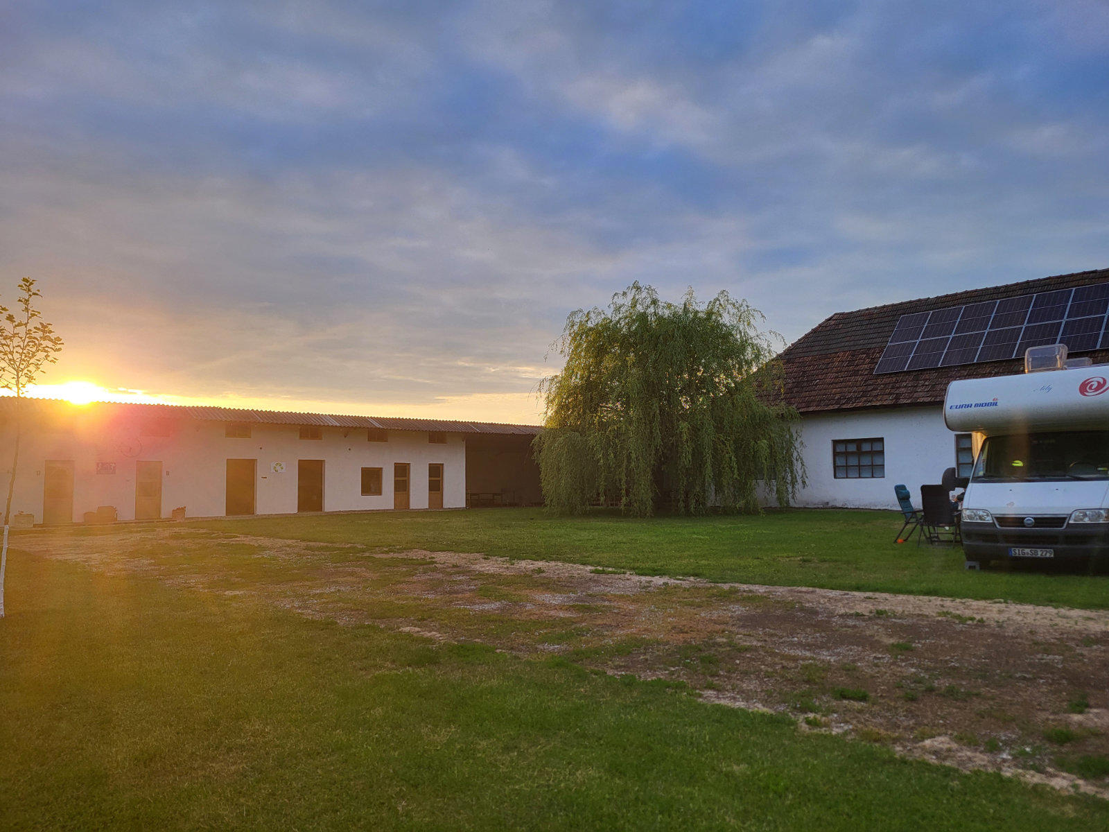 Zwei wei&szlig;e Geb&auml;ude, gr&uuml;ner Rasen mit unbefestigtem Weg, Weidebaum in der Mitte, Wohnmobil rechts, Sonne links am Horizont bei Sonnenuntergang, teilweise bew&ouml;lkter Himmel