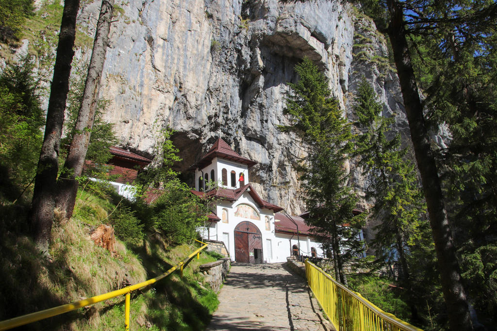 Wei&szlig;es Geb&auml;ude mit rotbraunen D&auml;chern und einer bogenf&ouml;rmigen Holzt&uuml;r an der Basis einer steilen Kalksteinwand; gepflasterter Weg mit gelbem Metallgel&auml;nder f&uuml;hrt darauf zu; Nadelb&auml;ume links und rechts