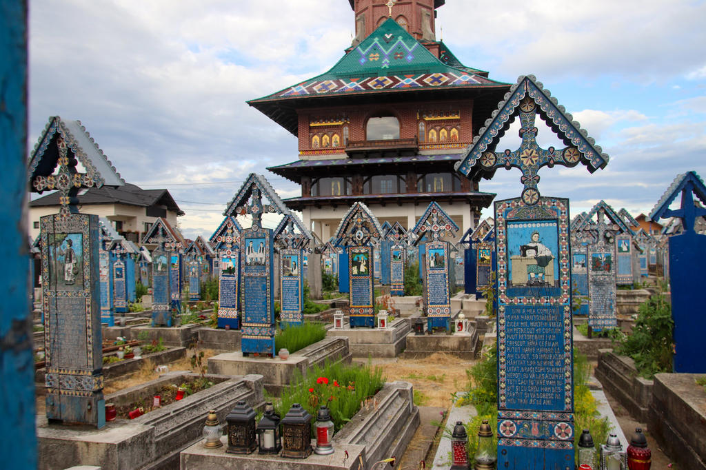 Friedhof mit Reihen bunt bemalter, geschnitzter h&ouml;lzerner Grabkreuze und Tafeln, Blumen und Laternen, h&ouml;lzerne Kirche mit Turm im Hintergrund, bew&ouml;lkter Himmel