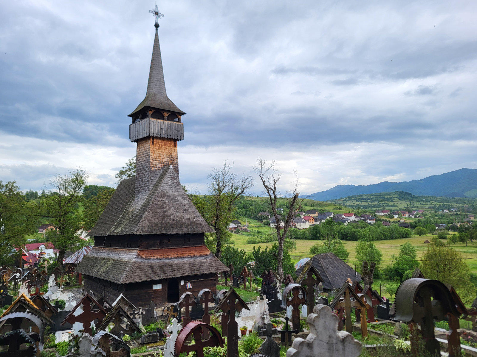Holzkirche mit steilem Schindeldach und hohem Turm, im Vordergrund zahlreiche Holzkreuze und Grabsteine, dahinter gr&uuml;ne Felder, H&auml;user und bew&ouml;lkter Himmel