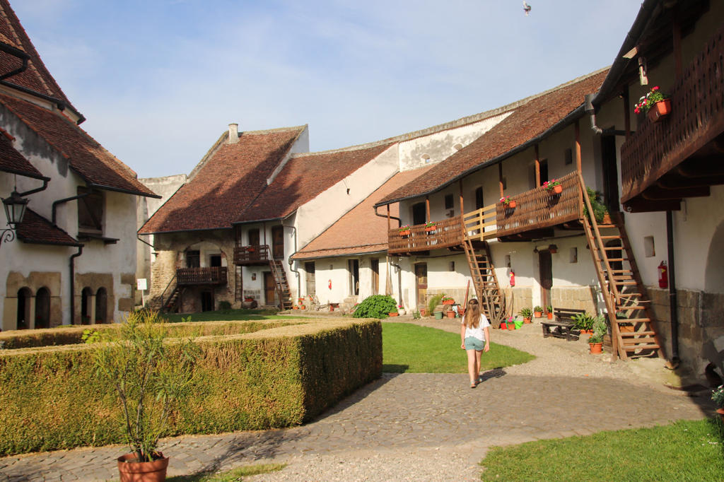 Innenhof mit wei&szlig; verputzten Geb&auml;uden, h&ouml;lzernen Balkonen und Treppen, steinigem Weg, gr&uuml;nem Rasen, geschnittener Hecke, Blument&ouml;pfen und einer Person in Shorts, die den Hof &uuml;berquert