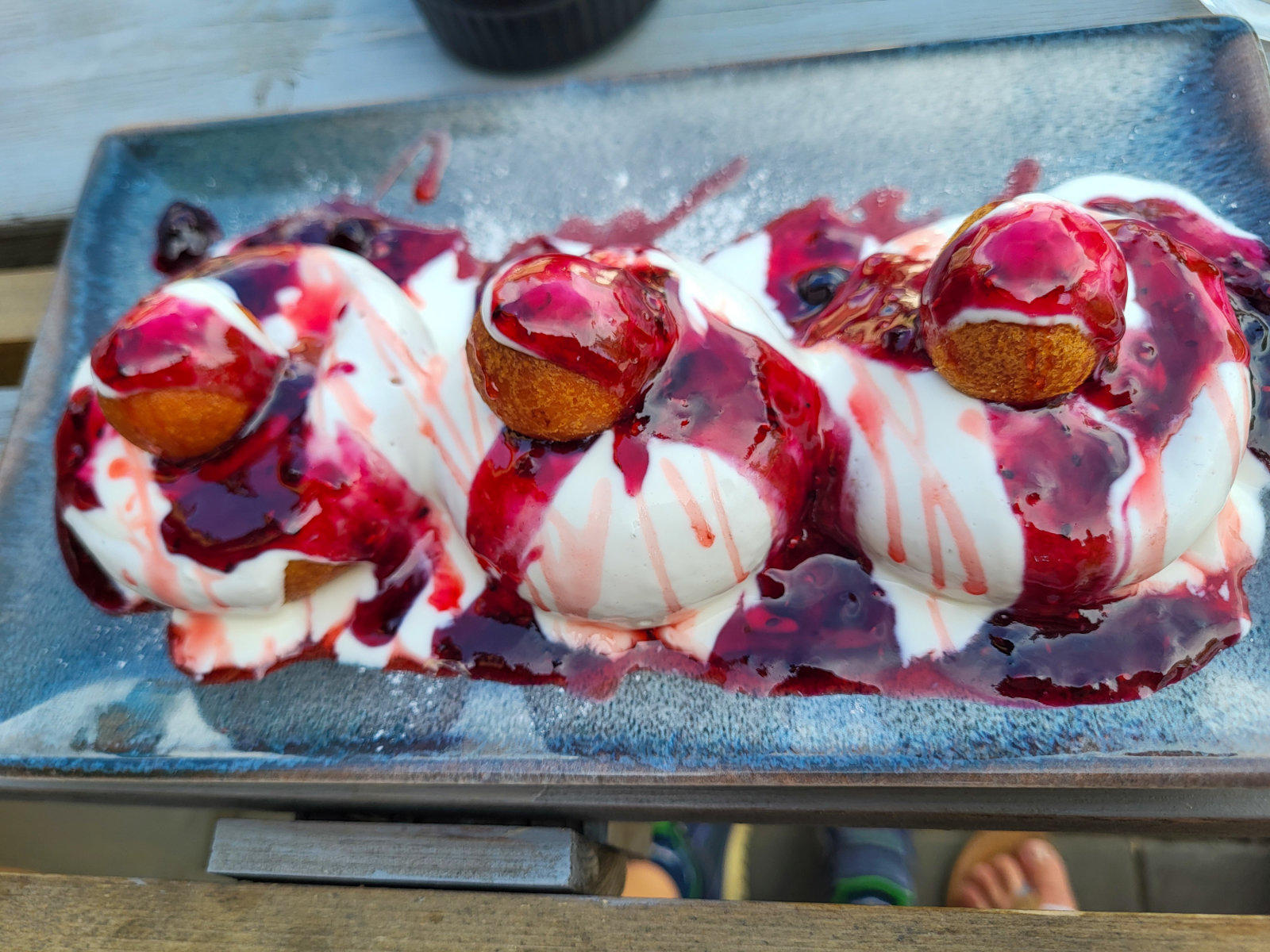 Drei wei&szlig;e Eiskugeln, jeweils mit einem kleinen braunen Geb&auml;ck belegt und mit dunkelroter Sauce &uuml;bergossen auf einem rechteckigen dunklen Teller; Holztisch und unten teilweise sichtbare F&uuml;&szlig;e mit Sandalen
