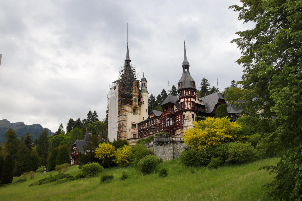 Gro&szlig;es steinernes Geb&auml;ude mit mehreren spitzen T&uuml;rmen, ein Turm mit Ger&uuml;st, gr&uuml;ne Wiese und B&auml;ume, bew&ouml;lkter Himmel