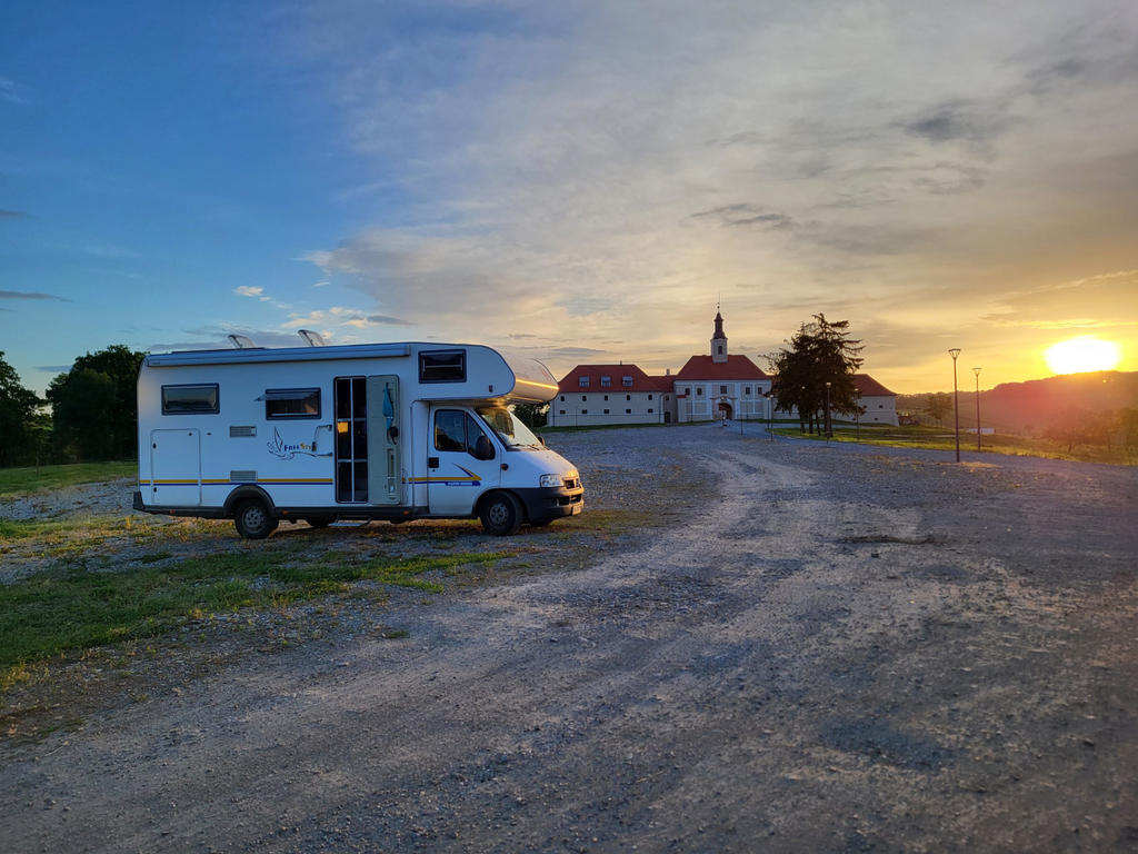 Wei&szlig;es Wohnmobil auf kiesigem Parkplatz, dahinter gro&szlig;es wei&szlig;es Geb&auml;ude mit rotem Dach und Turm, Sonne am Horizont, wolkiger Himmel