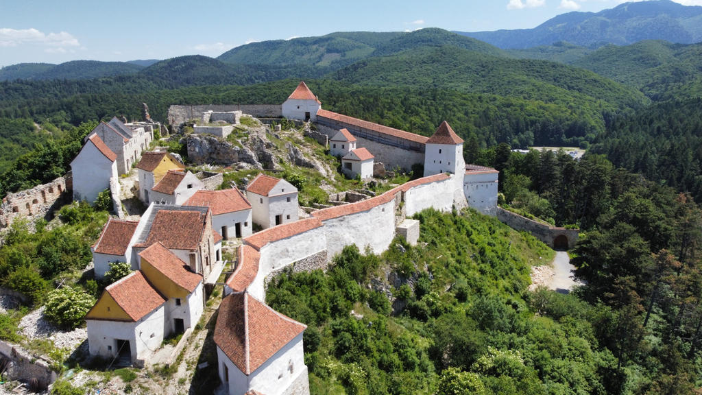 Lange wei&szlig;e Mauern und mehrere Geb&auml;ude mit roten Ziegeld&auml;chern auf einem bewachsenen H&uuml;gel; bewaldete H&auml;nge und Berge im Hintergrund; blauer Himmel mit Wolken