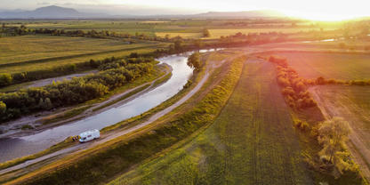 Luftaufnahme eines kurvenreichen Flusses durch Felder, ein wei&szlig;es Wohnmobil steht auf einem unbefestigten Uferweg, B&auml;ume am Fluss, tiefstehende Sonne rechts, entfernte Berge im Hintergrund