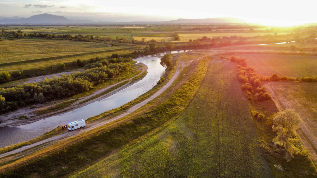 Luftaufnahme eines kurvenreichen Flusses durch Felder, ein wei&szlig;es Wohnmobil steht auf einem unbefestigten Uferweg, B&auml;ume am Fluss, tiefstehende Sonne rechts, entfernte Berge im Hintergrund