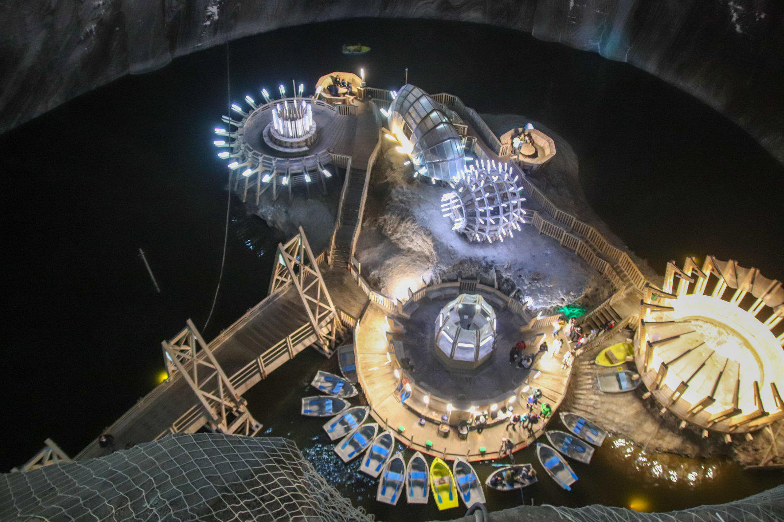 Gro&szlig;e unterirdische H&ouml;hle mit runden, beleuchteten Plattformen und Stegen um ein dunkles Wasserbecken, gelbe und blaue Ruderboote und eine Holzbr&uuml;cke