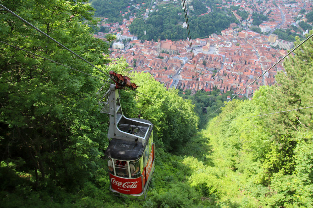 Kabine einer Seilbahn mit Coca-Cola-Logo &uuml;ber einem bewaldeten Hang, darunter Stadt mit roten Ziegeld&auml;chern