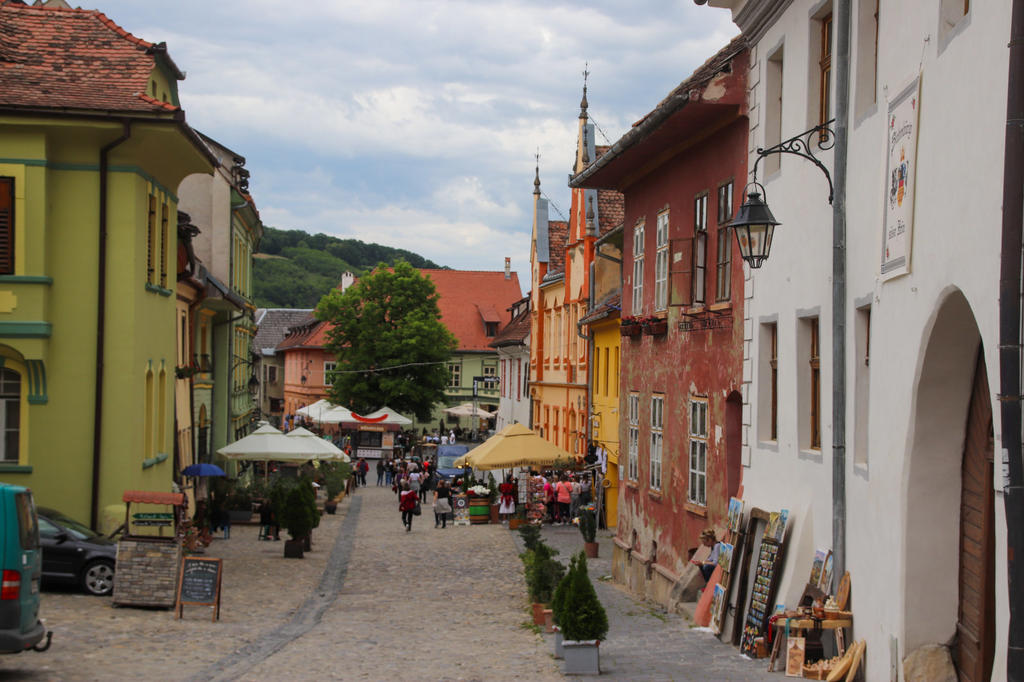 Kopfsteinpflasterstra&szlig;e mit bunten H&auml;usern, Fu&szlig;g&auml;ngern, Au&szlig;entischen mit Sonnenschirmen und H&uuml;geln im Hintergrund unter bew&ouml;lktem Himmel