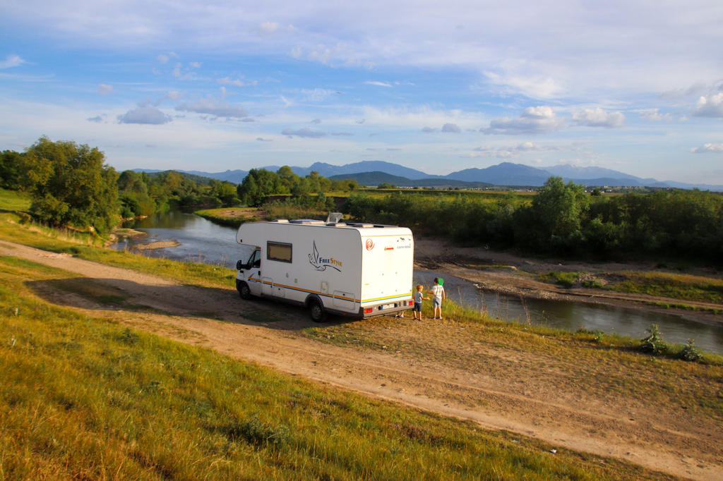 Wohnmobil auf einem unbefestigten Weg am Flussufer, zwei Personen stehen am Heck, B&auml;ume und Berge im Hintergrund, teils bew&ouml;lkter Himmel
