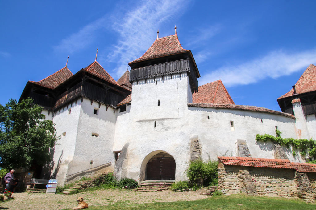 Wei&szlig;e Steinmauer mit Torbogen und mehreren T&uuml;rmen mit roten Ziegeld&auml;chern; blauer Himmel; Rasen; links sitzende Personen; vorn ein Hund