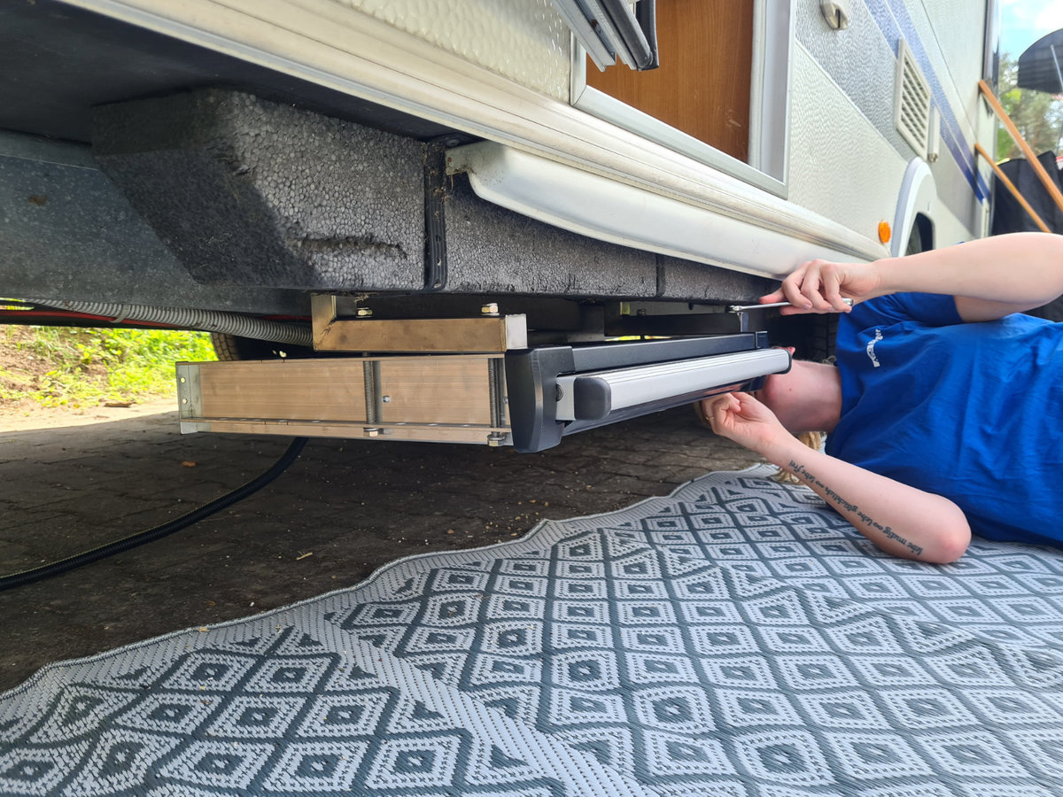 Unterseite eines Wohnmobils mit ausgezogener Metallstufe; zwei H&auml;nde halten die Stufe; Holzkl&ouml;tze und gemusterte Bodenmatte