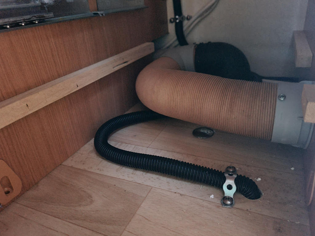 Holzfu&szlig;boden und -wand, beiger flexibler Schlauch an grauem Anschluss, schwarzer Wellschlauch auf dem Boden mit Metallschelle und Kunststoffhalter, rundes schwarzes Abdeckteil