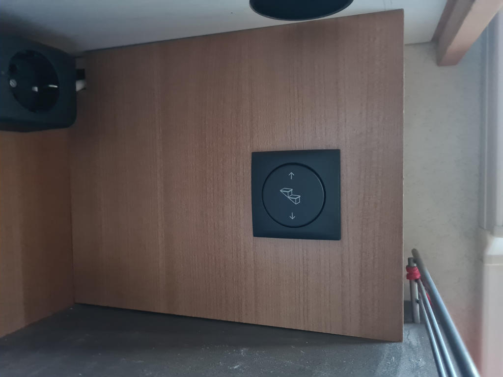 Holzverkleidung mit quadratischer schwarzer Platte mit rundem Symbol, rundes schwarzes Ger&auml;t oben links, Metallgel&auml;nder rechts mit roten Endkappen, heller Wandbereich rechts, dunkler Boden unten