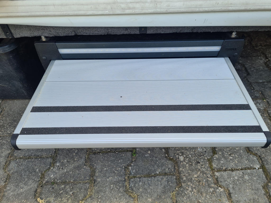 Wei&szlig;e Metallstufe mit zwei schwarzen Antirutschstreifen, dar&uuml;ber eine schwarze Halterung und die Unterkante einer Fahrzeugt&uuml;r; darunter graue Pflastersteine