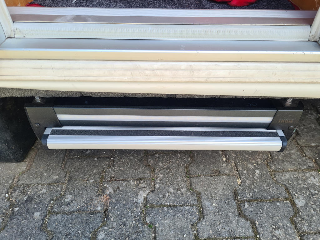 Wei&szlig;e T&uuml;r mit einem darunter montierten silbernen Metallbalken mit schwarzen Endkappen; graue Pflastersteine im Vordergrund; roter Teppich im Innenbereich sichtbar