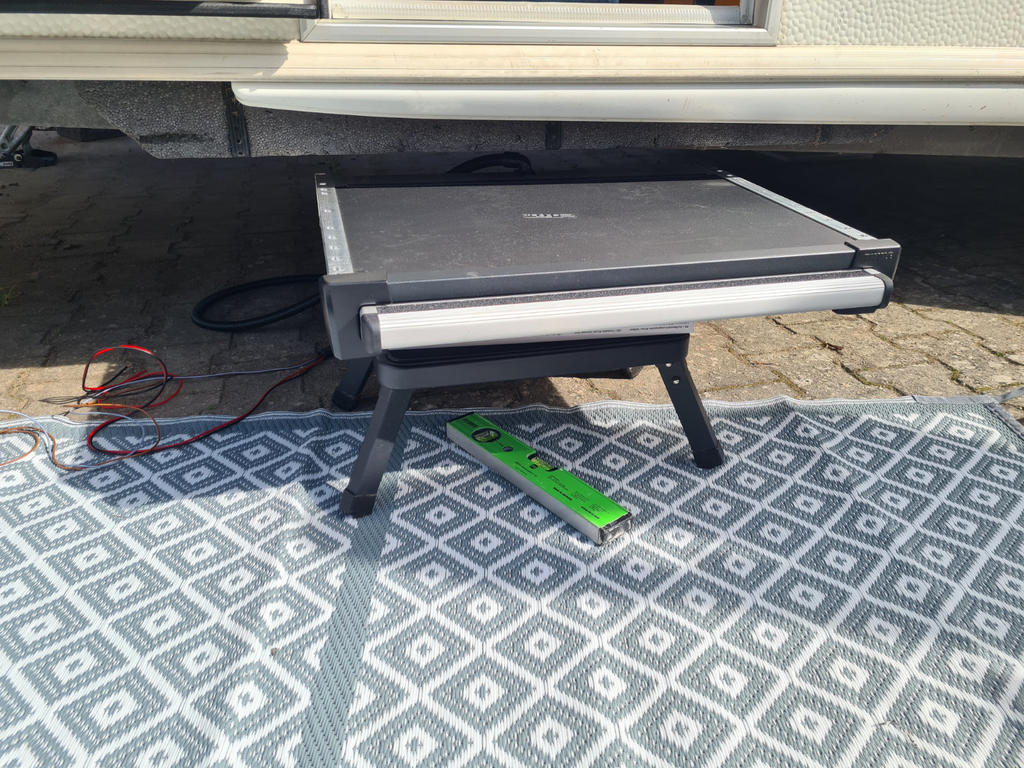 Ausgeklappte Metallstufe mit schwarzer Oberfl&auml;che und silbernem Rand auf zwei Beinen unter einem Fahrzeug, auf grauem, gemustertem Outdoor-Teppich; gr&uuml;ne Wasserwaage auf dem Teppich; rote und schwarze Kabel links; Pflastersteine rechts