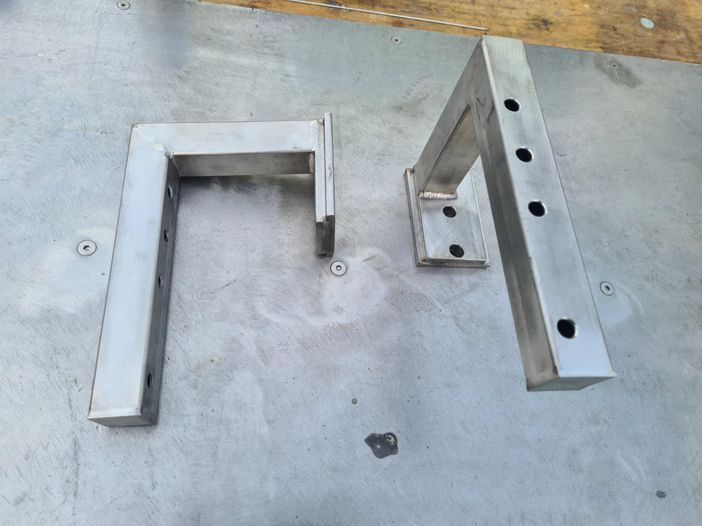 Zwei L‑f&ouml;rmige Edelstahlwinkel auf grauer Metallfl&auml;che, sichtbare Schraubenk&ouml;pfe und Holzkante oben