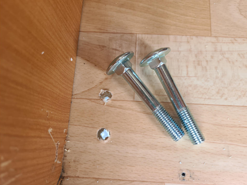 Zwei silberne Schrauben liegen auf einem hellen Holzfu&szlig;boden neben zwei Muttern in Bohrl&ouml;chern; links ist eine Holzkante sichtbar