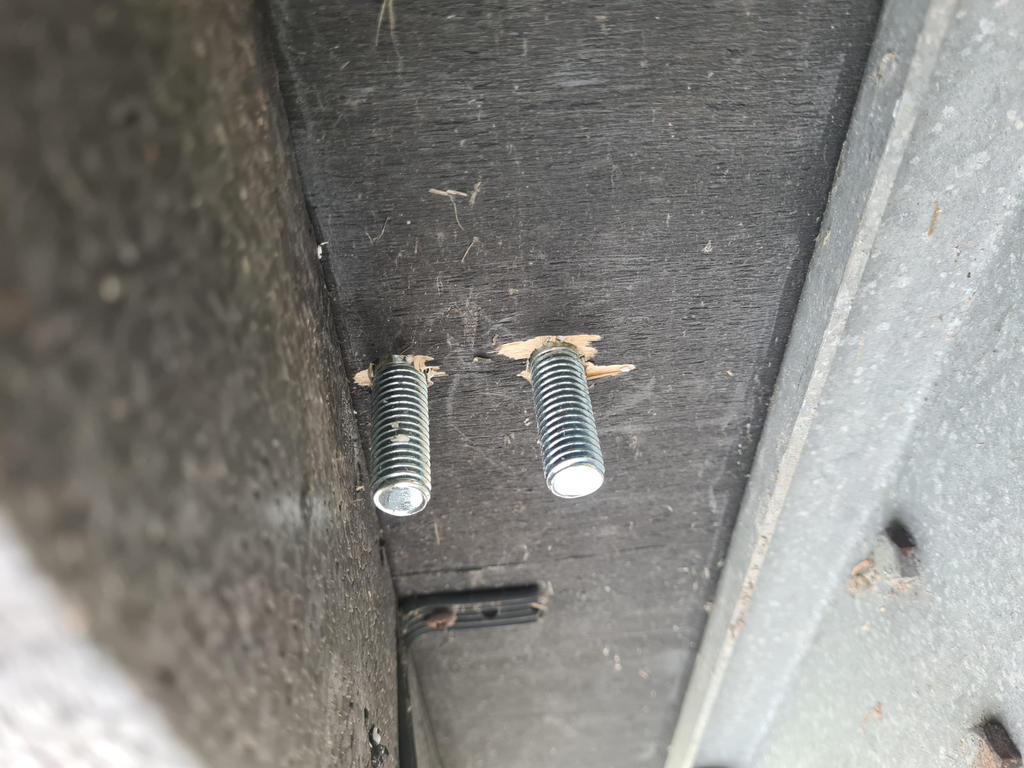 Zwei Metallgewindebolzen ragen aus einem dunklen Holzbrett, Holzsplitter um die L&ouml;cher, rechts ein grauer Metallrahmen sichtbar