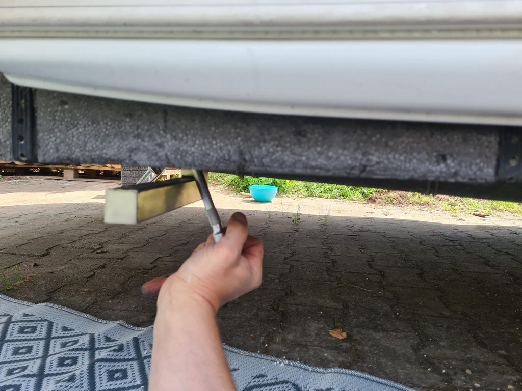 Hand mit Schraubenschl&uuml;ssel unter einem Fahrzeug; Matte auf dem Pflaster, Kunststoffblock und gr&uuml;ner Eimer im Hintergrund