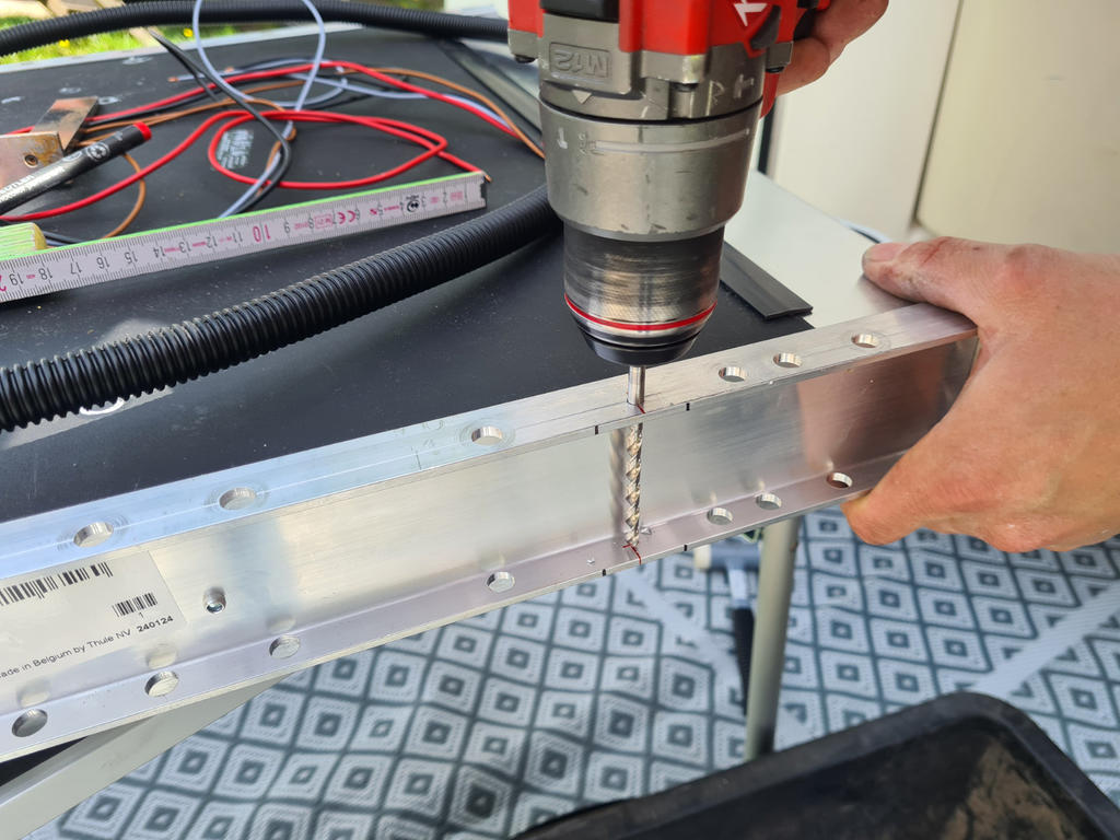 Zwei H&auml;nde halten einen Aluminiumrahmen, eine rote Handbohrmaschine mit Spiralbohrer ber&uuml;hrt den Rahmen; im Hintergrund Kabel, ein Ma&szlig;band und eine schwarze Wanne auf einer gemusterten Matte