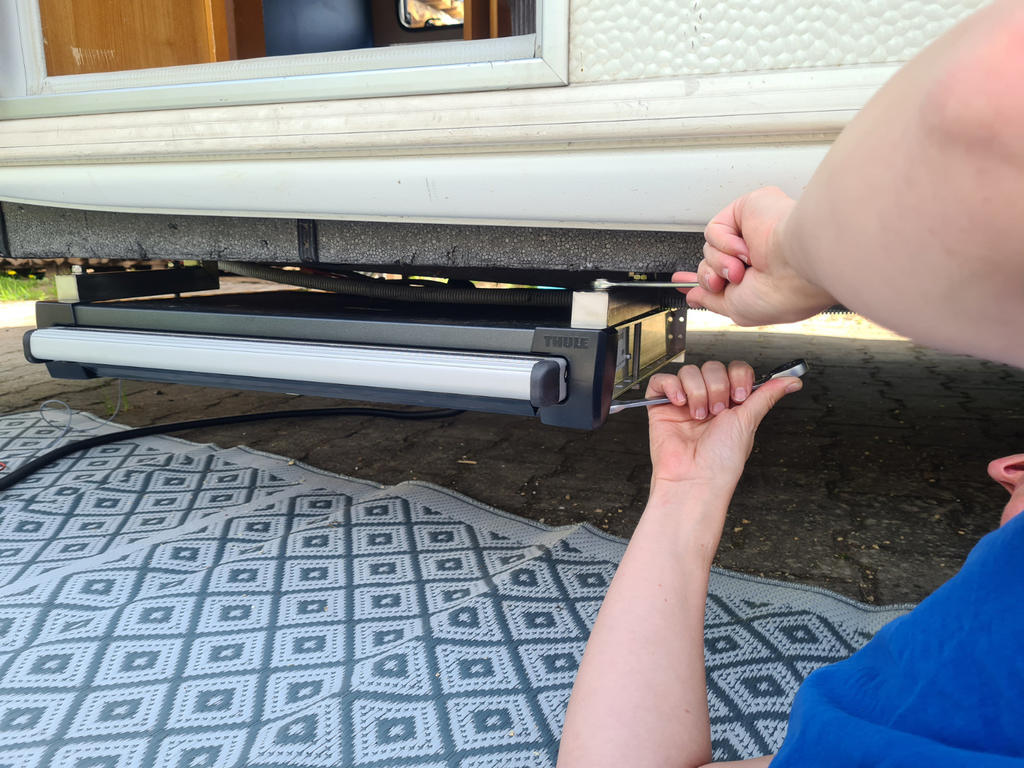 H&auml;nde einer Person unter einem Wohnwagen halten Schraube und Werkzeug an einer Metallhalterung mit l&auml;nglicher Leuchte; gemusterte Matte auf dem Boden, wei&szlig;e Au&szlig;enwand des Wohnwagens mit Fenster