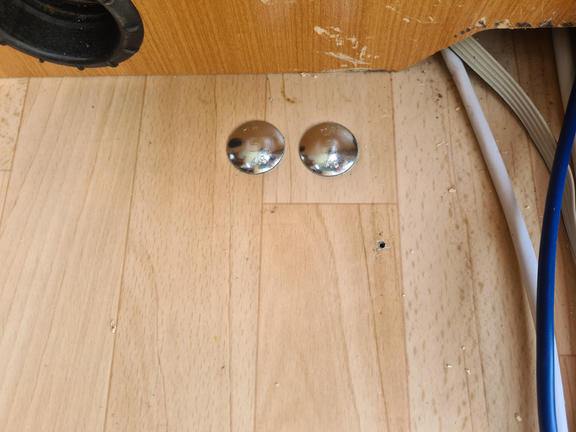 Heller Holzfu&szlig;boden mit zwei runden, silbernen Metallabdeckungen nebeneinander; links oben Schrankunterseite mit rundem Ausschnitt; rechts blaue und graue Kabel