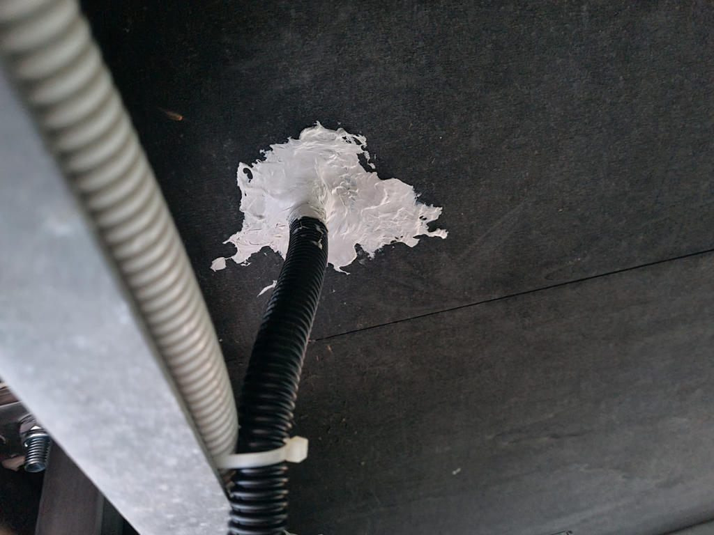 Dunkles Flachpaneel mit wei&szlig;er Dichtmasse um ein schwarzes, gewelltes Kabel; links ein graues Wellrohr mit wei&szlig;em Kabelbinder