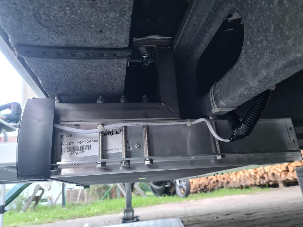 Unterseite mit horizontalem Metalltr&auml;ger, daran befestigte rechteckige Metallbox mit sichtbaren Schrauben und f&uuml;hrenden Kabeln; schwarzer Wellschlauch, graue D&auml;mmung oben, St&uuml;tzbein, Gras und Holzstapel im Hintergrund