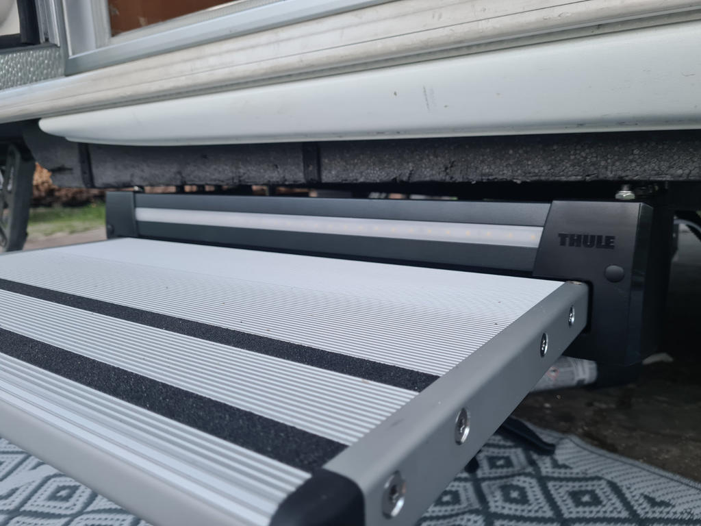 Ausgeklappte zweiteilige Aluminium-Trittstufen mit schwarzen Anti-Rutsch-Streifen unter einer Fahrzeugschwelle, seitliches Geh&auml;use mit Schriftzug Thule