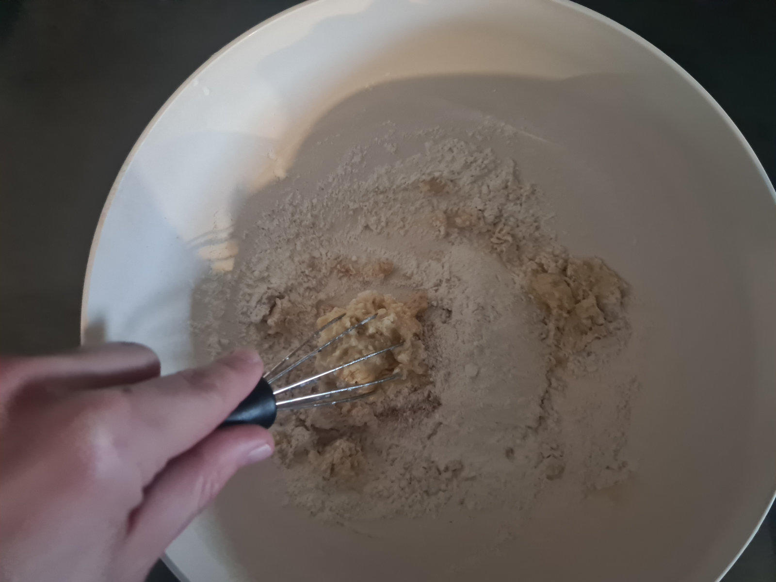 Hand h&auml;lt kleinen Schneebesen &uuml;ber einer wei&szlig;en Sch&uuml;ssel mit hellem Pulver und Kr&uuml;meln