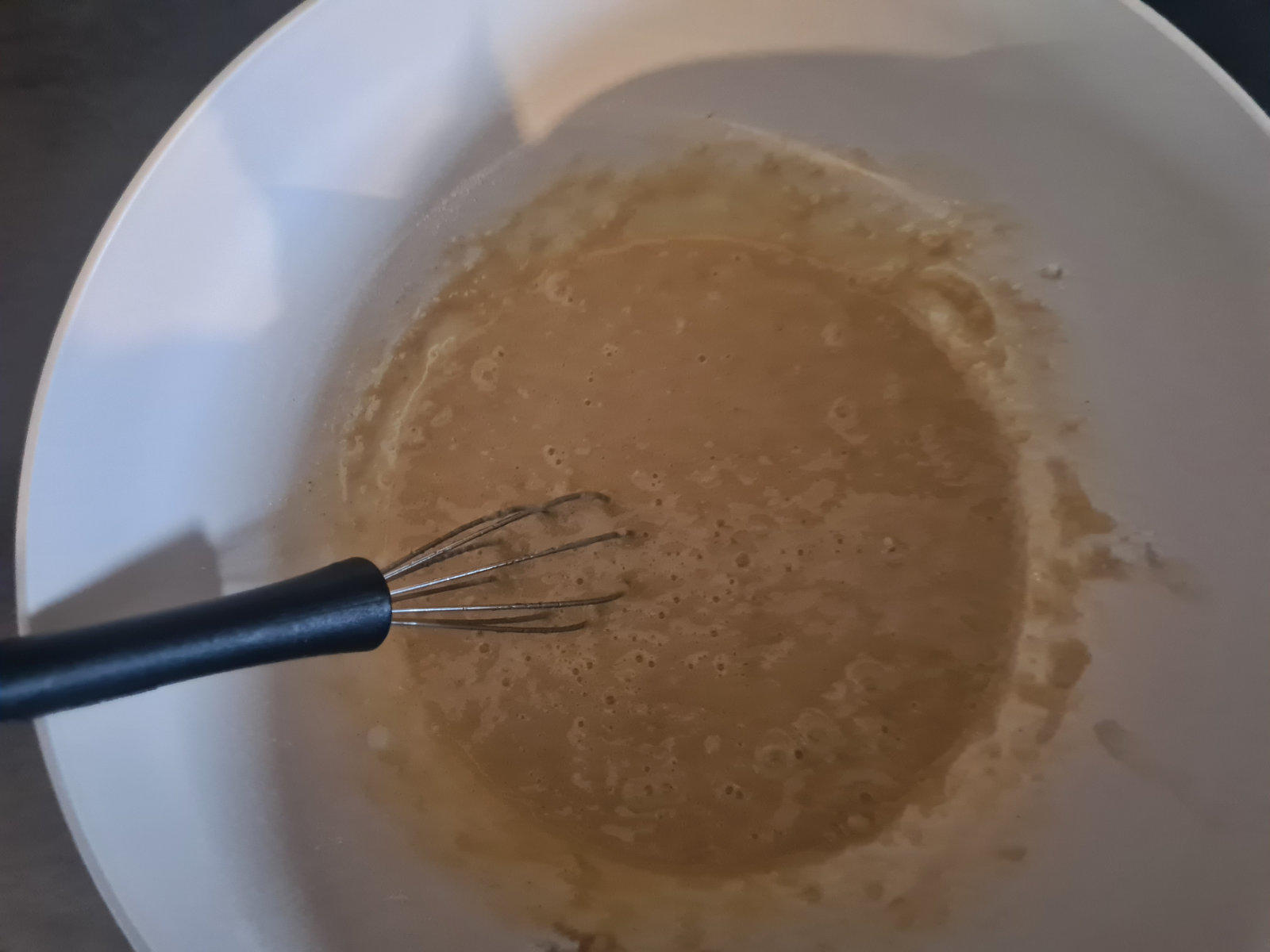 Wei&szlig;e Sch&uuml;ssel mit beigefarbener, fl&uuml;ssiger Masse und kleinem Schneebesen mit schwarzem Griff