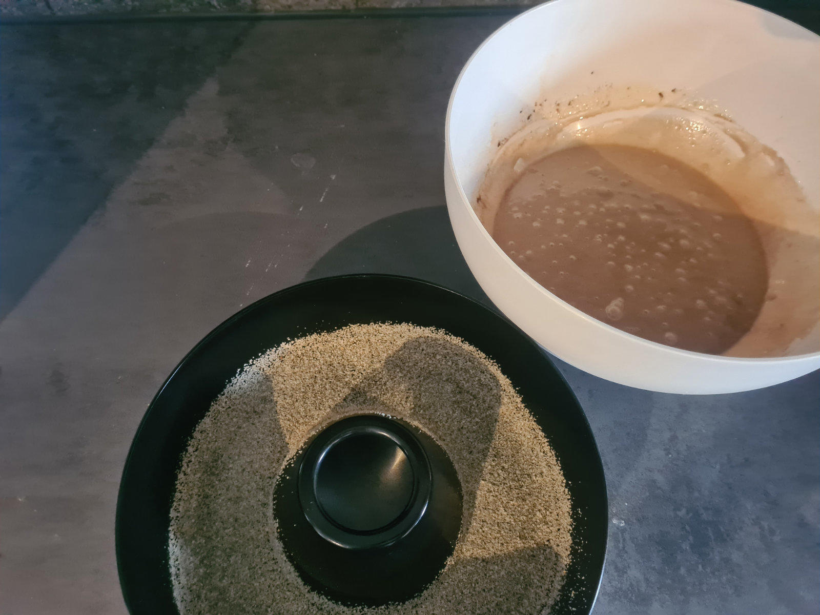 Wei&szlig;e Sch&uuml;ssel rechts mit hellbeiger Fl&uuml;ssigkeit und Schaumresten; links schwarzer runder Teller mit hellbeigem, feinem, ringf&ouml;rmig angeordnetem Material und kleinem schwarzen runden Objekt in der Mitte; graue Unterlage