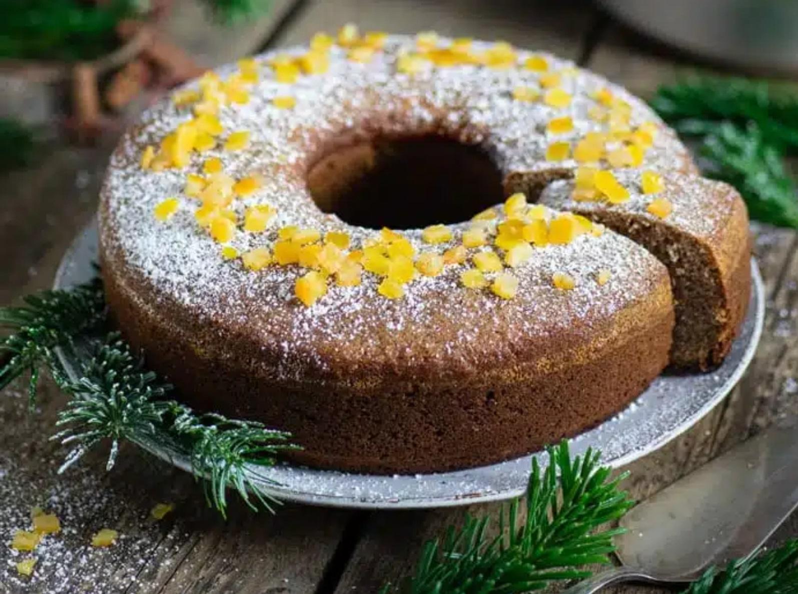 Ringf&ouml;rmiger Kuchen mit einem fehlenden St&uuml;ck, bestreut mit Puderzucker und belegt mit gelben Bl&uuml;tenbl&auml;ttern, auf einem Teller, umgeben von gr&uuml;nen Nadelzweigen