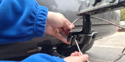 Zwei H&auml;nde mit blauem &Auml;rmel halten einen Schraubendreher und verbinden elektrische Kabel an einer Anh&auml;ngerkupplung unter der Sto&szlig;stange eines Autos