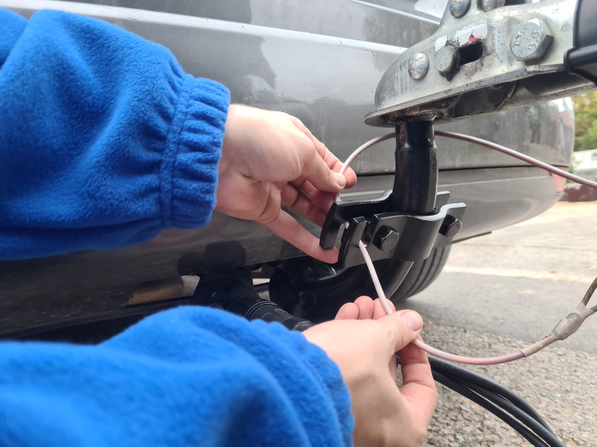 Zwei H&auml;nde mit blauem &Auml;rmel halten einen Schraubendreher und verbinden elektrische Kabel an einer Anh&auml;ngerkupplung unter der Sto&szlig;stange eines Autos