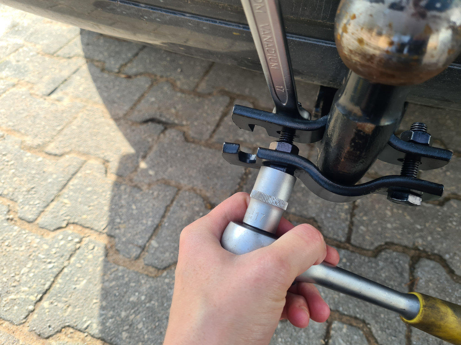 Hand h&auml;lt Steckschl&uuml;ssel an der Mutter der Anh&auml;ngerkupplungskugel unter einem Auto; Anh&auml;ngerkupplungskugel, Steckschl&uuml;ssel mit gelbem Griff und Pflastersteine sichtbar